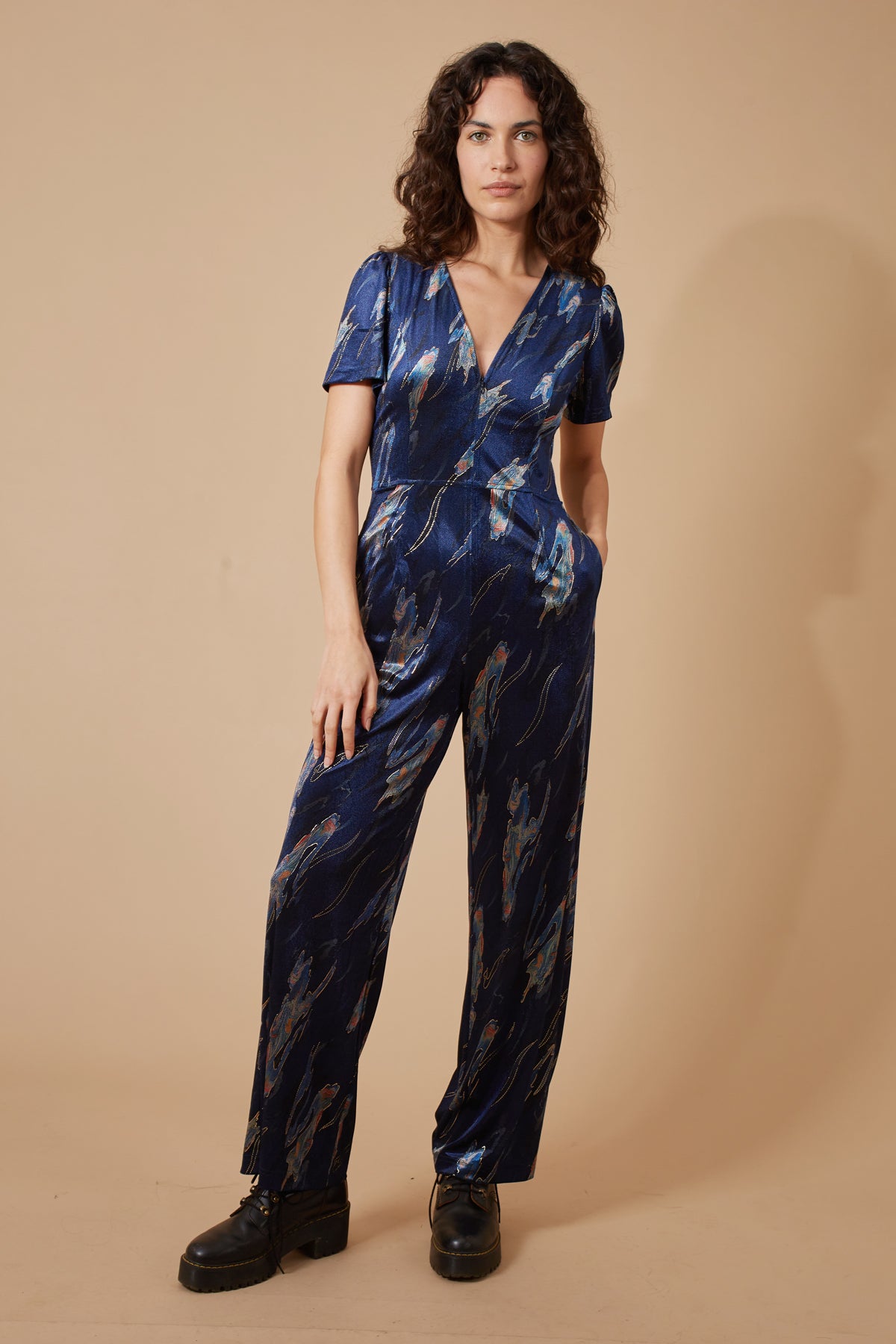 The beautiful Corrie jumpsuit from British brand TRAFFIC PEOPLE is a cracker for an event and to stand out from the crowd. The dark navy fabric has a multi coloured print and accents of gold throughout. The elegant V neck has a zip to the waist. There are short sleeves and side pockets. The leg is a straight shape so very flattering.